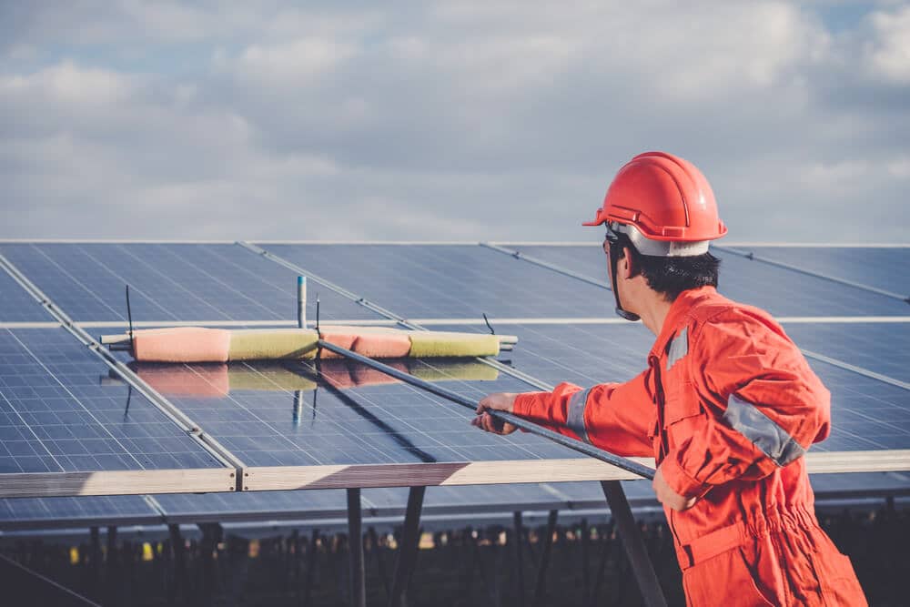 Man in red uniform cleaning solar panels manually with long handle stick with microfibre